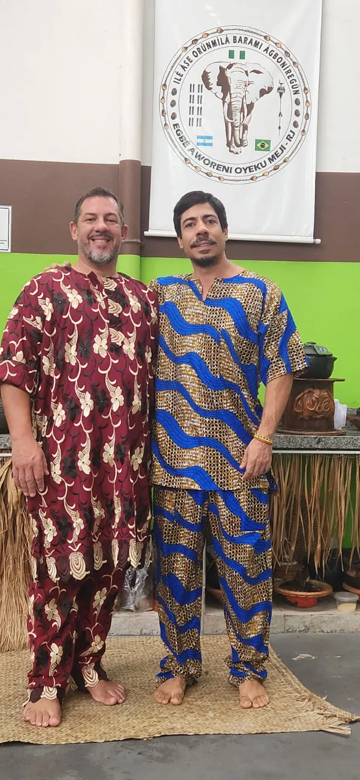 Pai Bruno, à direita, e seu iniciador no culto tradicional, de trajes yorubás e descalços sobre uma esteira, diante de um estandarte com a figura de um elefante