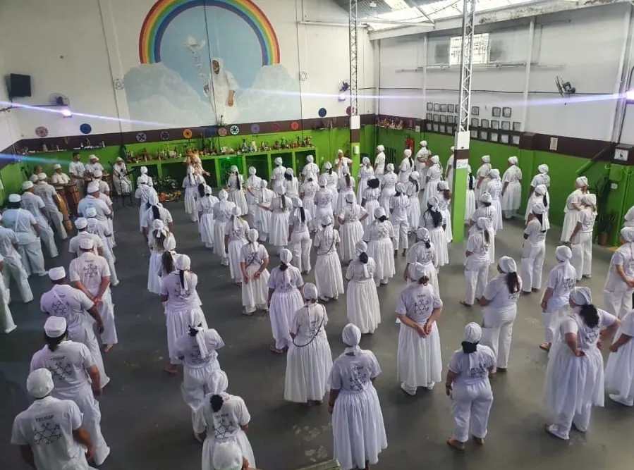 Médiuns do TEUSP vestidos de branco, em fileiras no salão do templo, diante do congá e de um mural com arco-íris