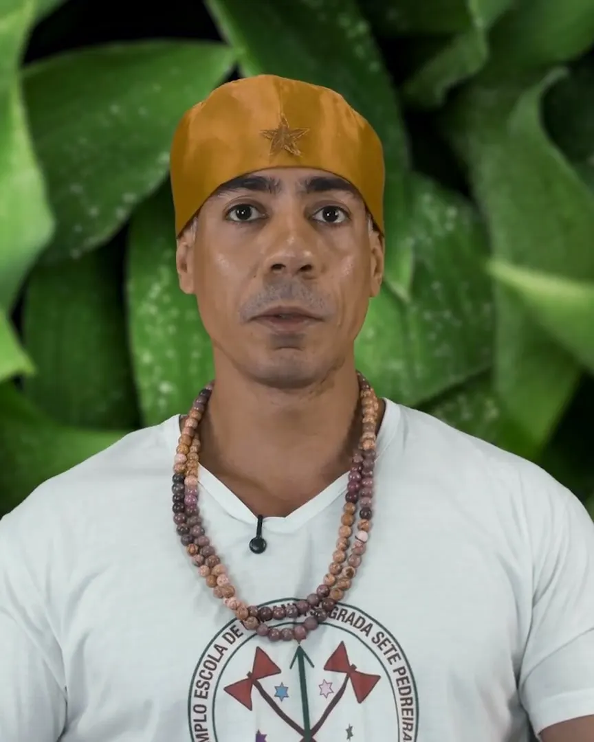 Bruno Amorim, de camiseta branca do templo, guias de contas e gorro dourado, diante de folhas verdes