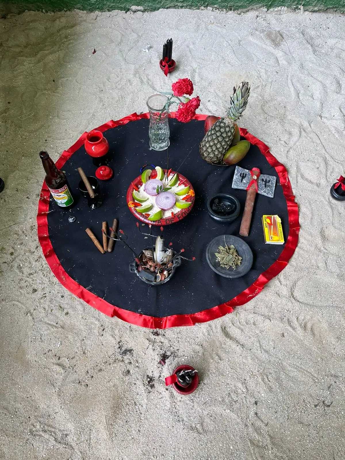 Oferenda sobre toalha preta com babado vermelho, na areia, com abacaxi, cerveja, charutos, caixa de fósforos e um prato de cebolas e pimentas