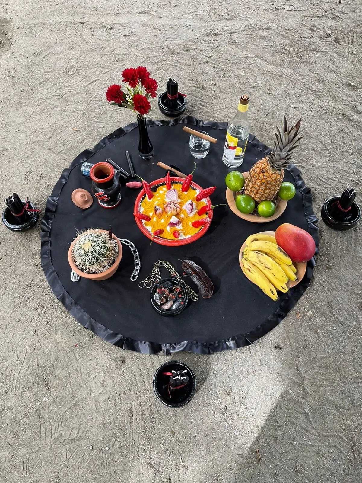 Oferenda a Exu sobre toalha preta com um cacto no vaso, corrente, abacaxi, bananas, manga, cravos vermelhos e velas em alguidares