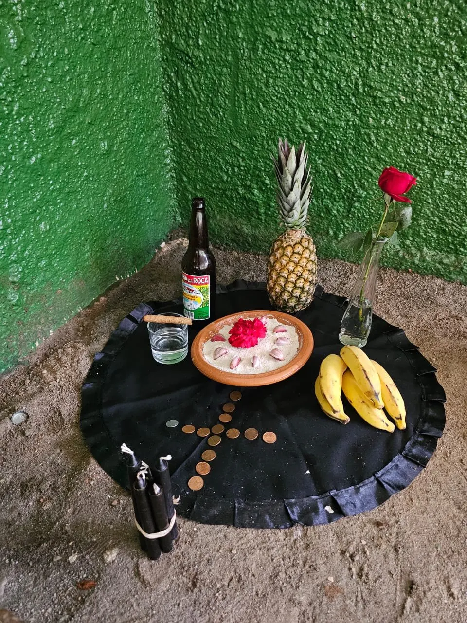 Oferenda a Exu no canto de uma parede verde, sobre toalha preta, com abacaxi, bananas, cerveja, uma rosa vermelha e uma cruz de moedas