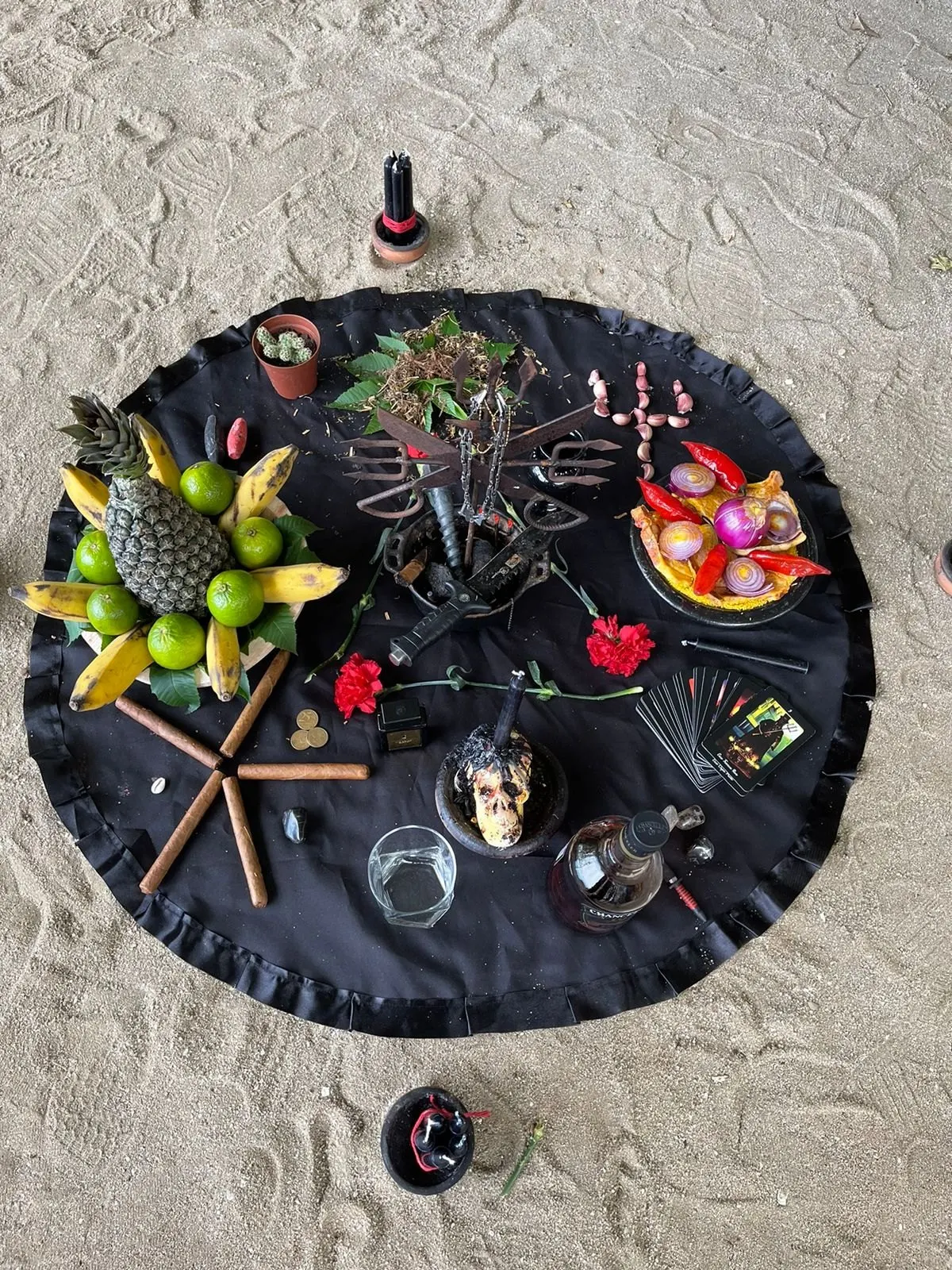 Oferenda sobre toalha preta na areia, com tridente de ferro, pequena caveira, charutos dispostos em estrela, abacaxi, bananas, limões, cravos vermelhos e pimentas