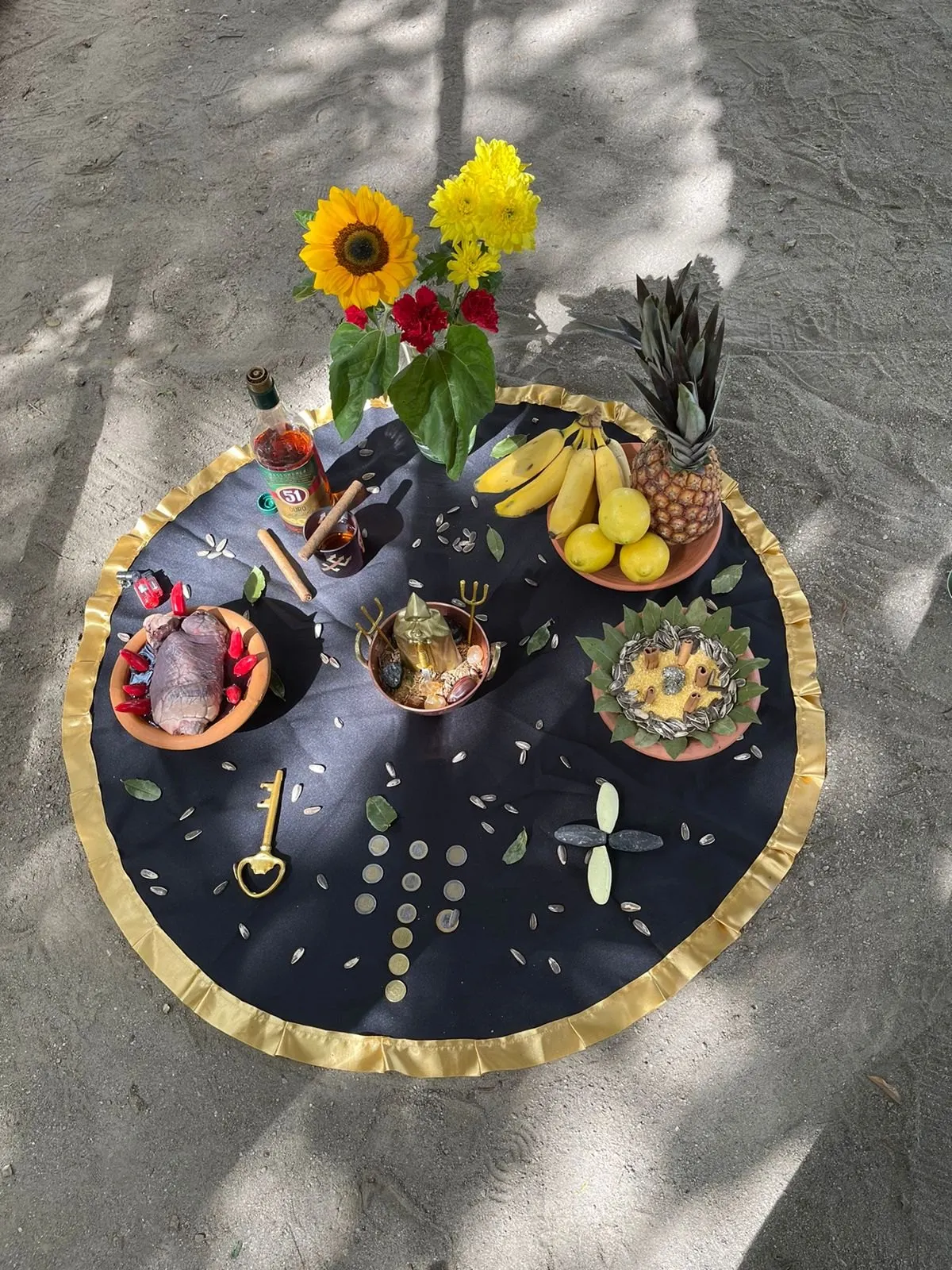 Oferenda sobre toalha preta com babado dourado, com girassol, bananas, abacaxi, limões e uma cruz de moedas