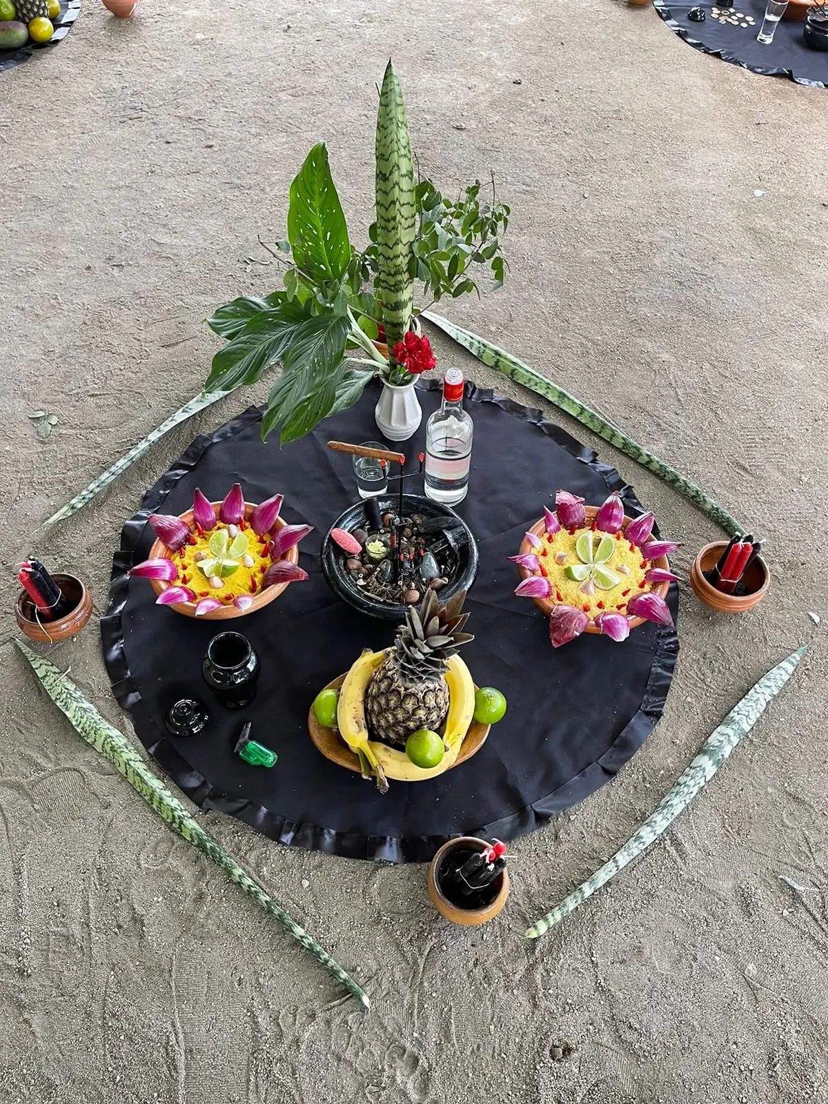 Oferenda a Exu sobre toalha preta na areia, cercada de folhas de espada-de-são-jorge, com pratos de frutas, abacaxi, cachaça e velas em alguidares