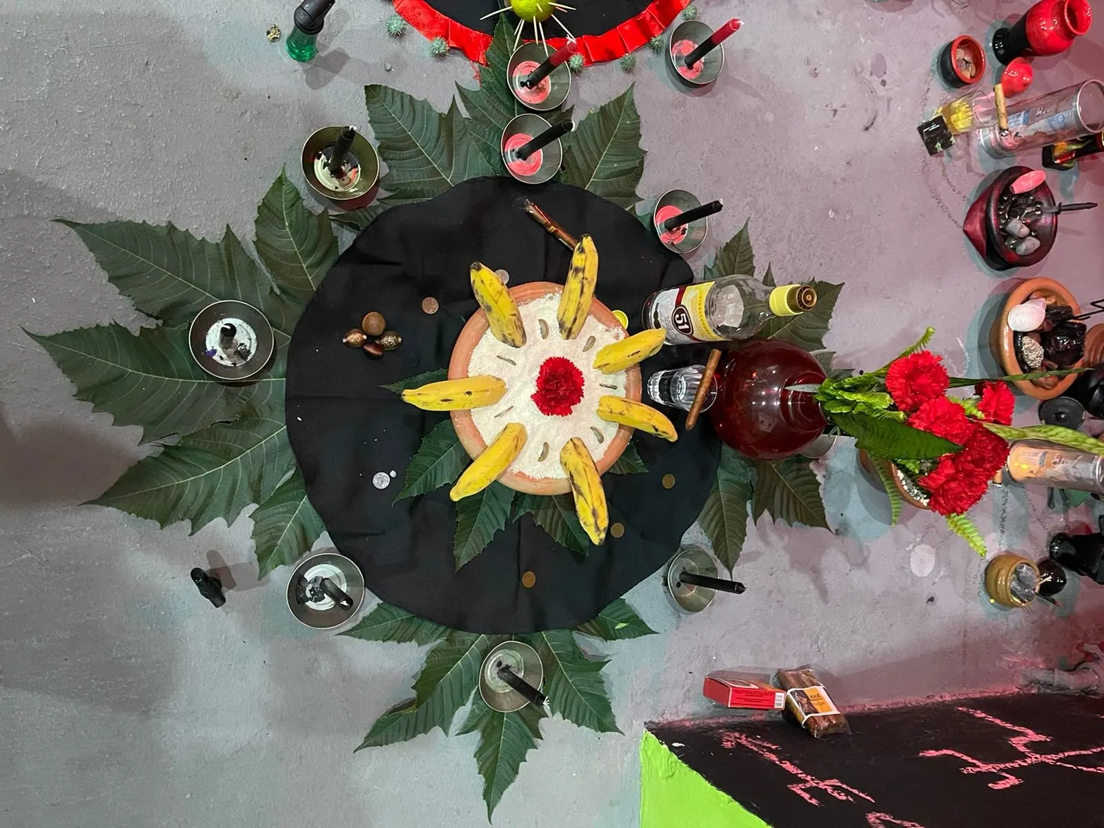 Oferenda a Exu vista de cima, sobre toalha preta cercada de folhas verdes, com bananas, flor vermelha e velas em alguidares