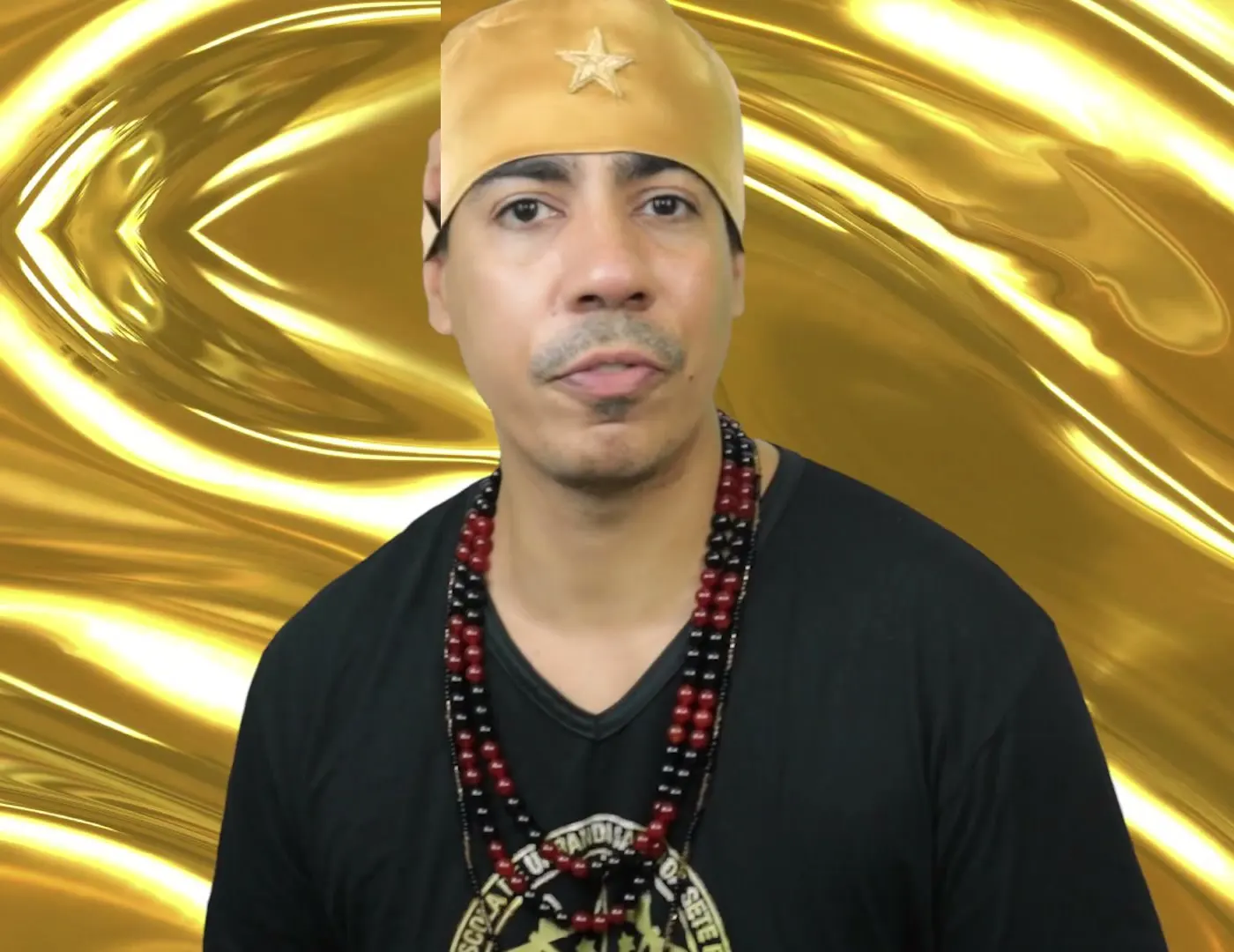 Bruno Amorim, de camiseta preta, guias vermelhas e pretas e gorro dourado, diante de um fundo dourado