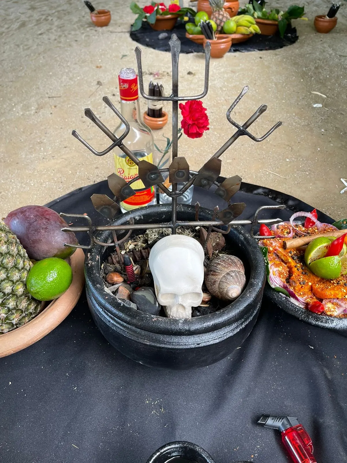 Tridentes de ferro cravados num alguidar preto com uma caveira branca e pedras, sobre toalha preta, entre pratos de frutas e pimentas
