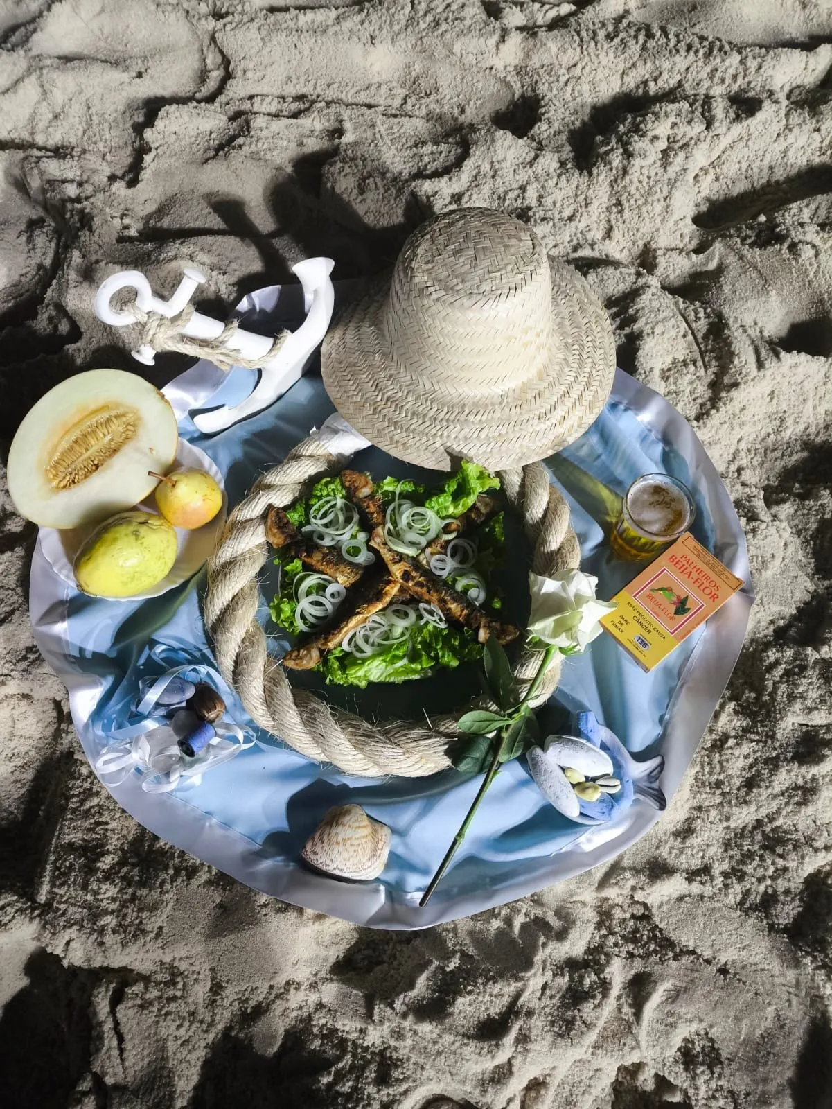 Oferenda vista de cima na areia, sobre tecido azul-claro, com chapéu de palha, âncora branca, corda, melão, peras e um prato de peixe sobre folhas