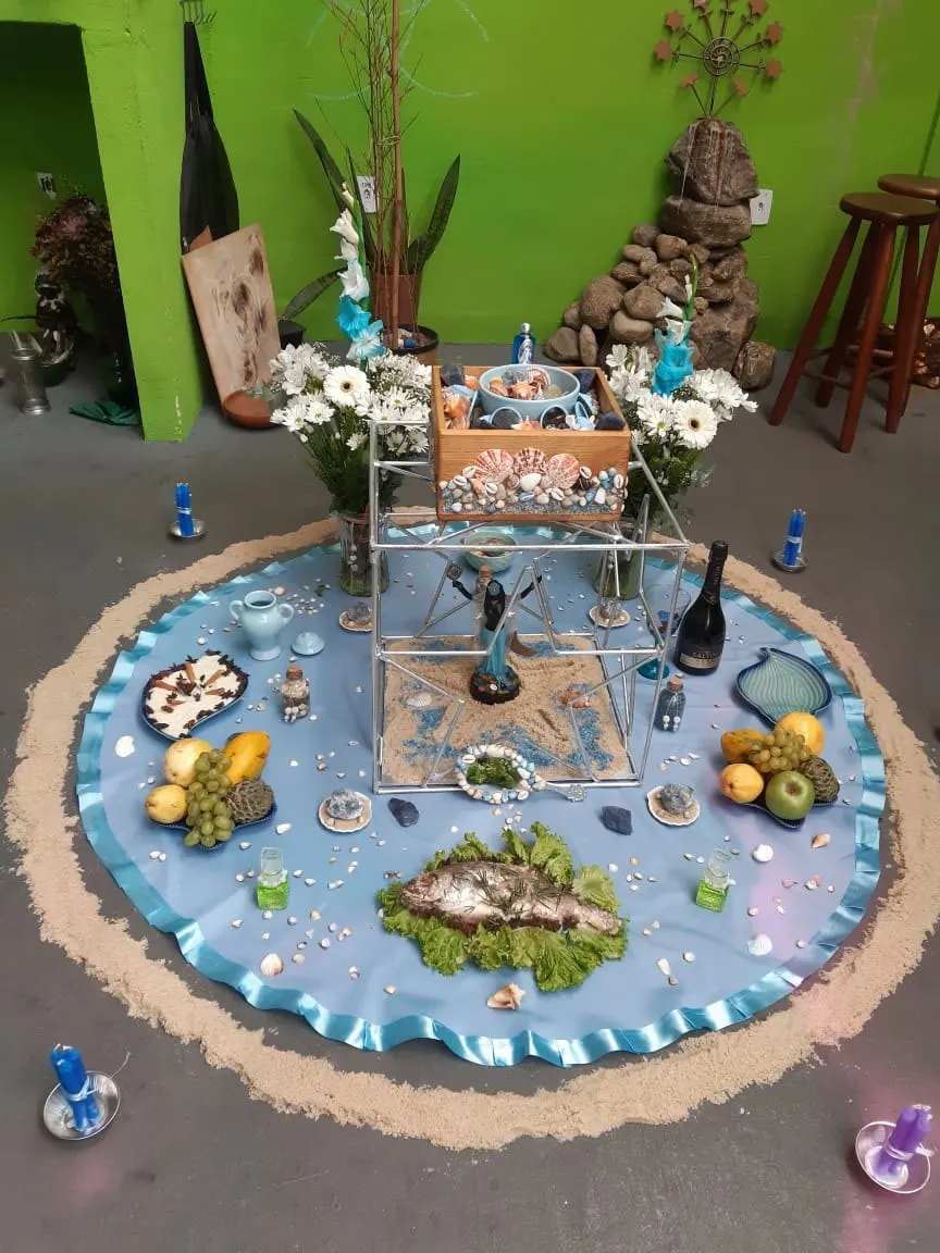 Oferenda a Iemanjá no chão do templo, sobre toalha azul-clara cercada de areia, com um peixe sobre folhas, conchas, frutas, flores brancas e velas azuis