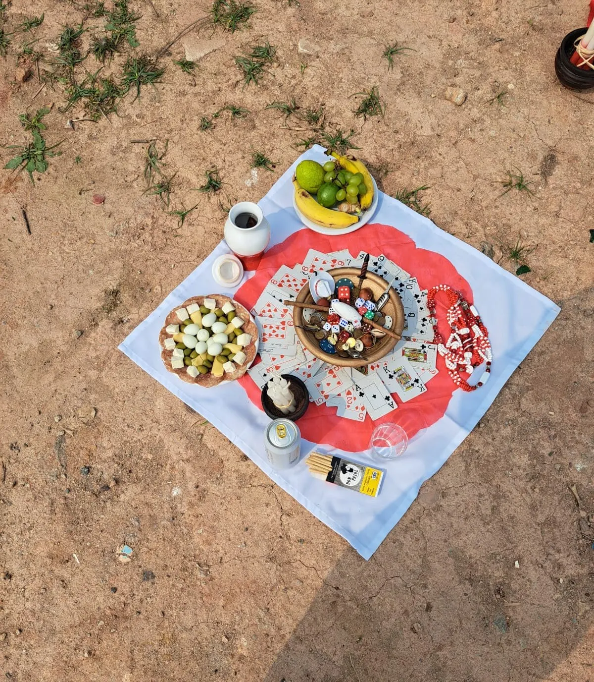 Oferenda vista de cima sobre toalha branca com círculo vermelho, com baralho, dados, ovos de codorna, frutas e uma caneca
