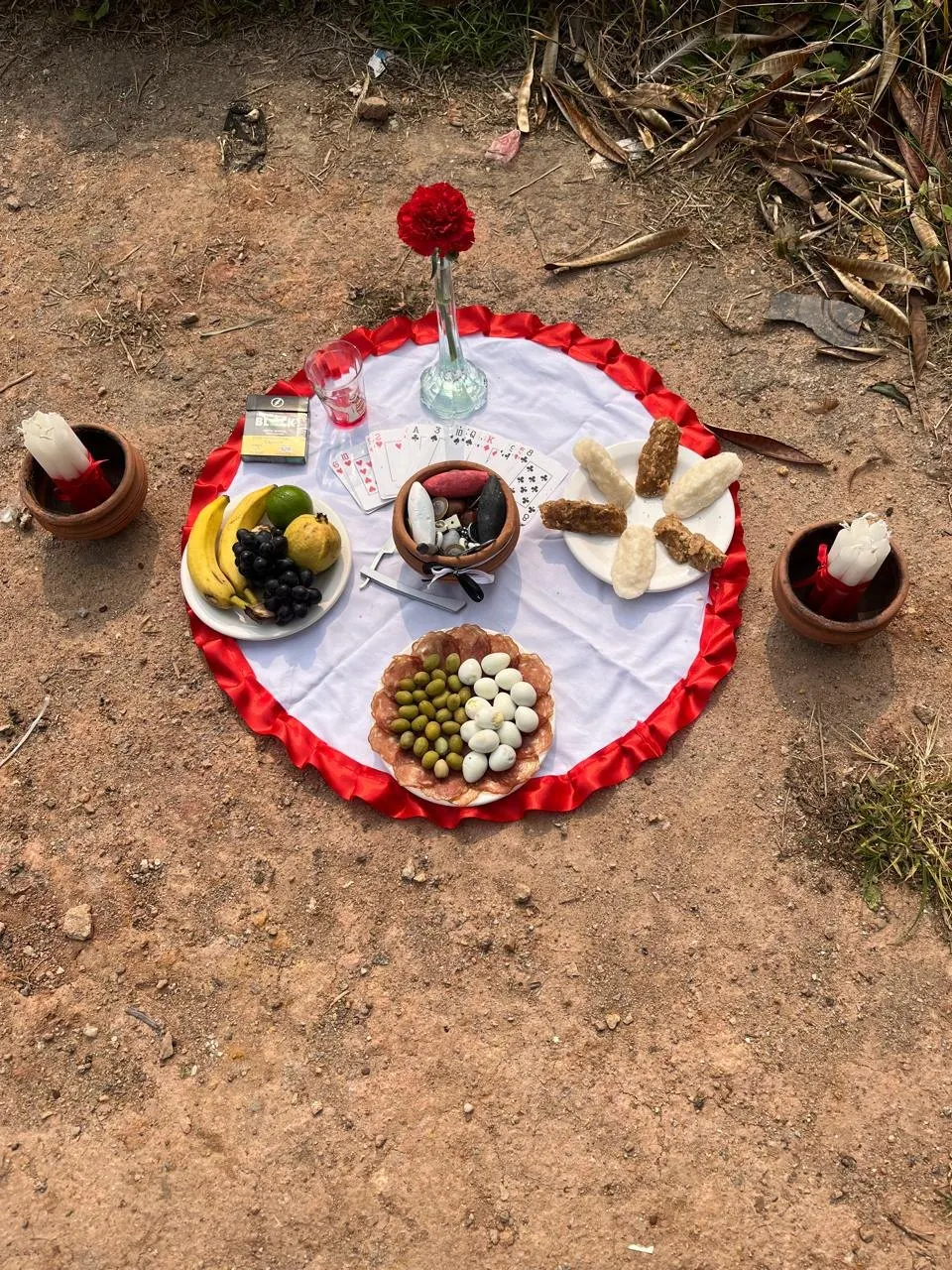 Oferenda sobre toalha branca com babado vermelho, na terra, com um cravo vermelho, baralho, frutas, ovos de codorna, azeitonas e velas em alguidares