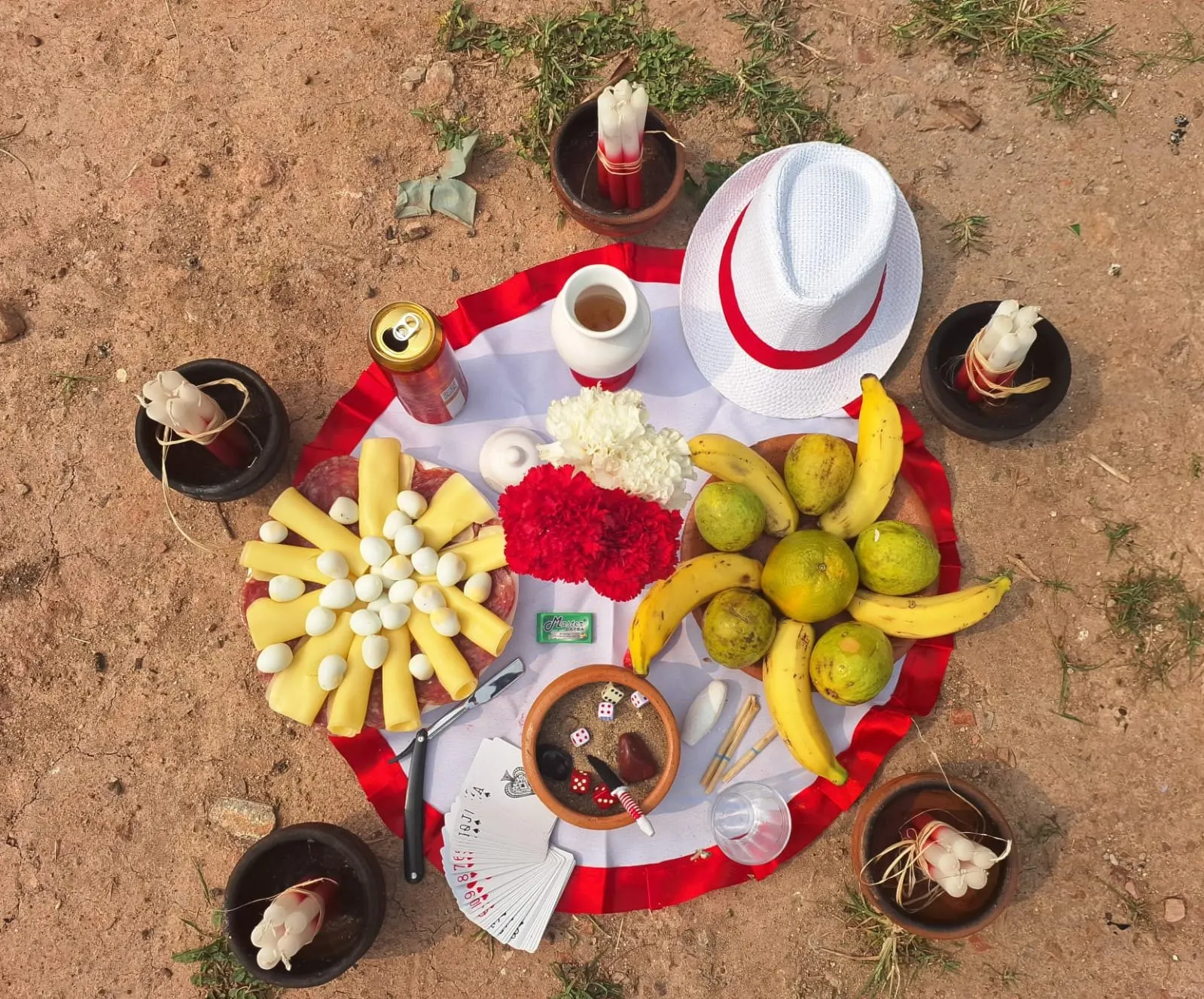 Oferenda vista de cima sobre toalha branca com babado vermelho, com chapéu branco, queijo, ovos de codorna, cravos vermelhos e brancos, bananas, laranjas, dados e baralho