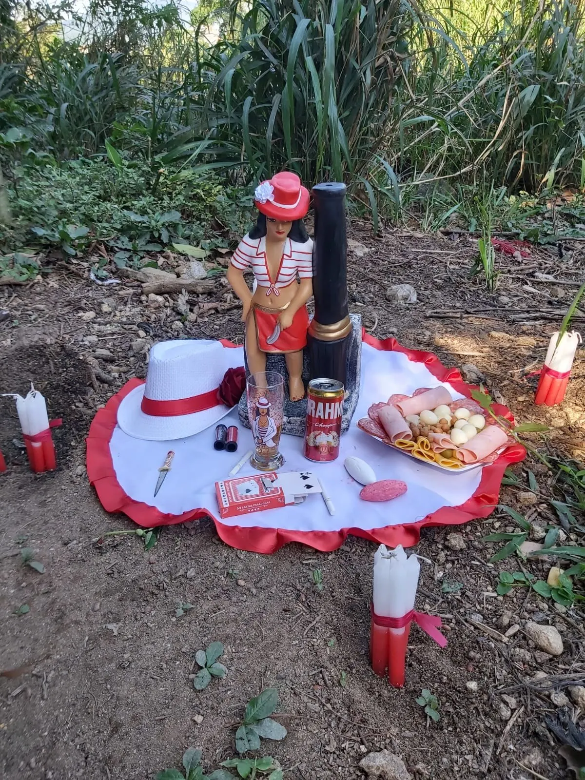 Imagem de chapéu vermelho e camisa listrada sobre toalha branca com babado vermelho, na mata, com chapéu branco, frios, cerveja, baralho e velas