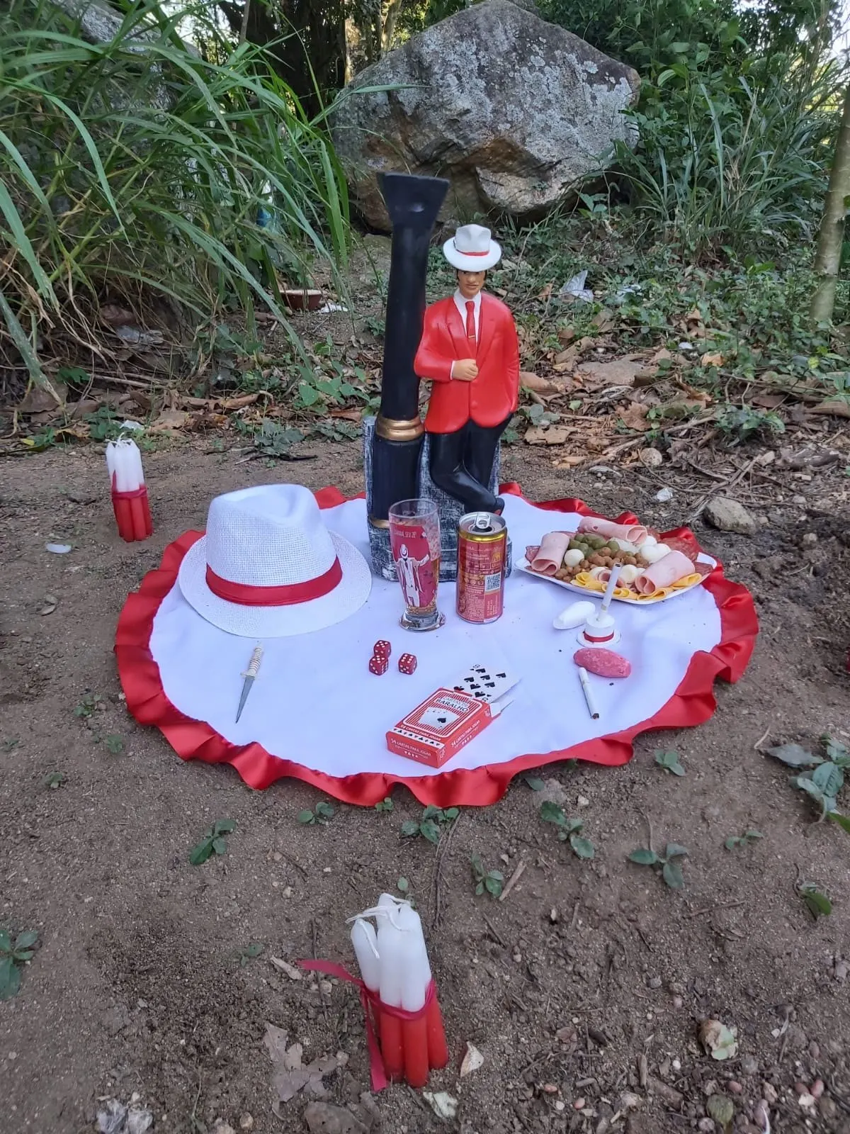 Imagem de Malandro de terno vermelho sobre toalha branca com babado vermelho, na mata, com chapéu branco, cerveja, petiscos, baralho e velas brancas e vermelhas