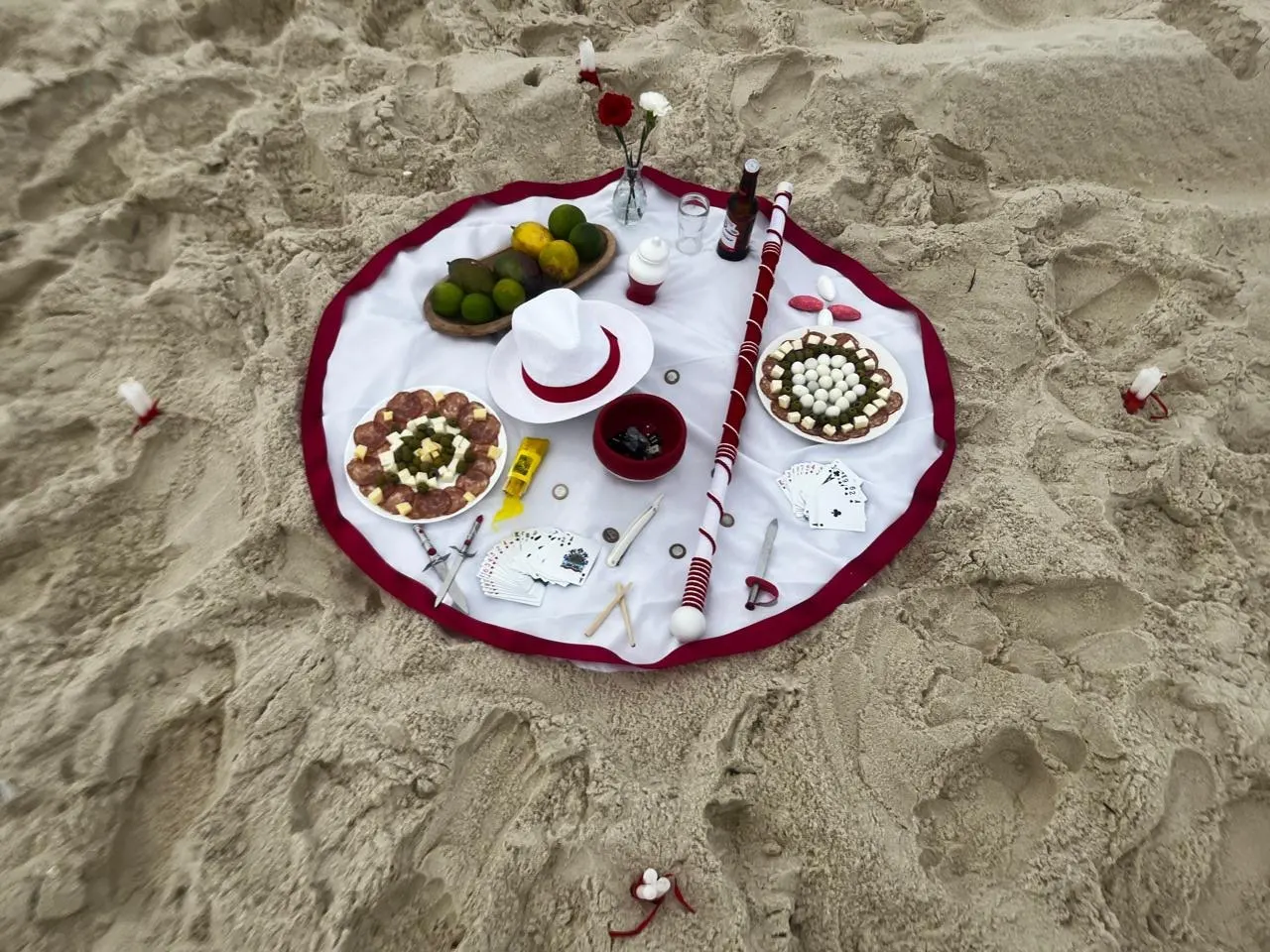 Oferenda vista de cima na areia da praia, sobre toalha branca com babado vinho, com chapéu branco, bengala, frutas, petiscos e cartas de baralho