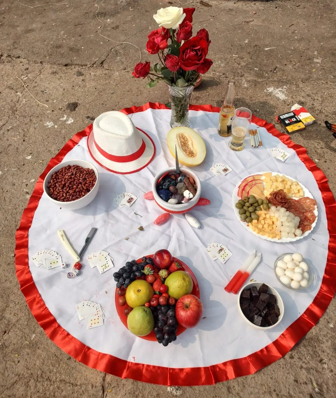 Oferenda sobre toalha branca com babado vermelho, com rosas, chapéu branco, melão, frios, amendoim, chocolate, frutas e cartas de baralho