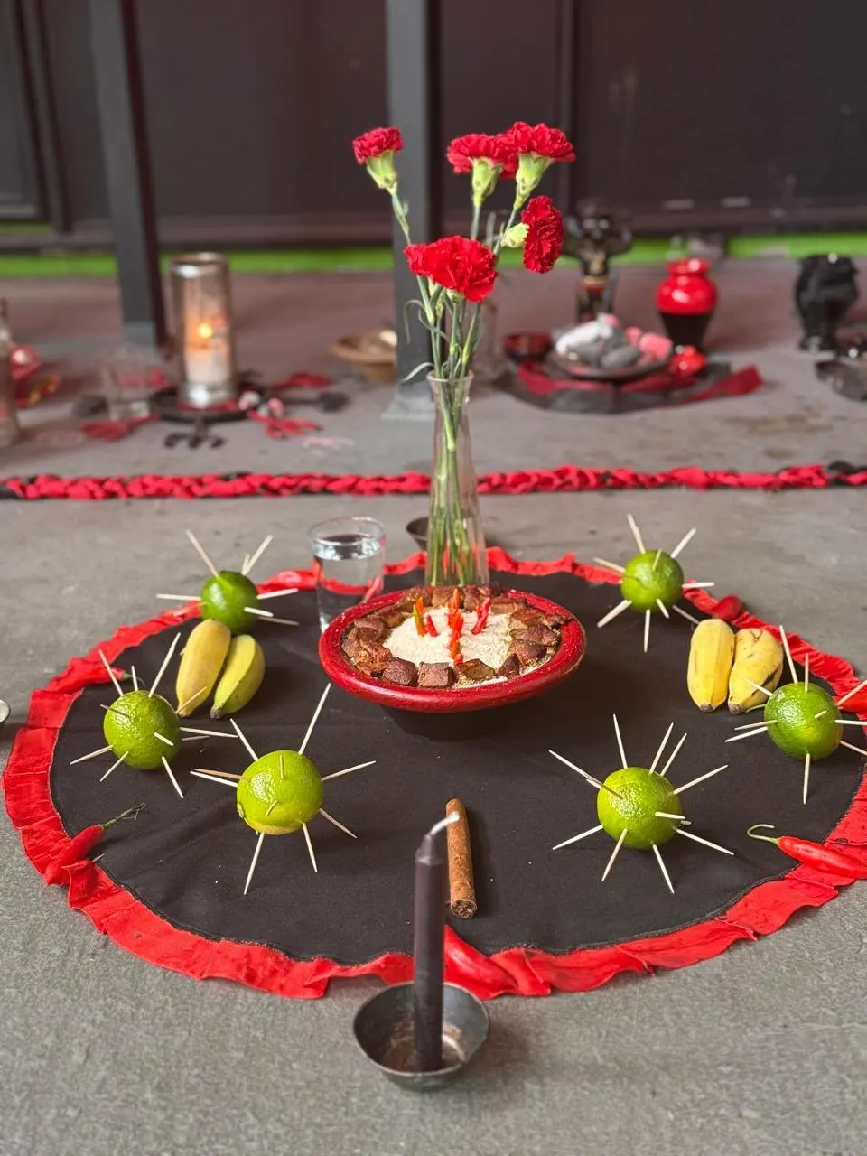 Oferenda sobre toalha preta com babado vermelho, com cravos vermelhos num vaso, limões espetados com palitos, bananas e uma vela escura