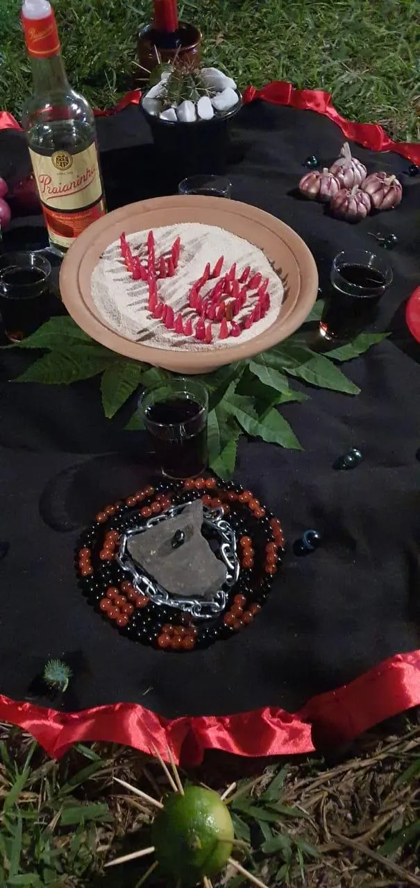 Detalhe de oferenda com prato de farofa e pimentas em espiral, garrafa e guia de contas sobre toalha preta