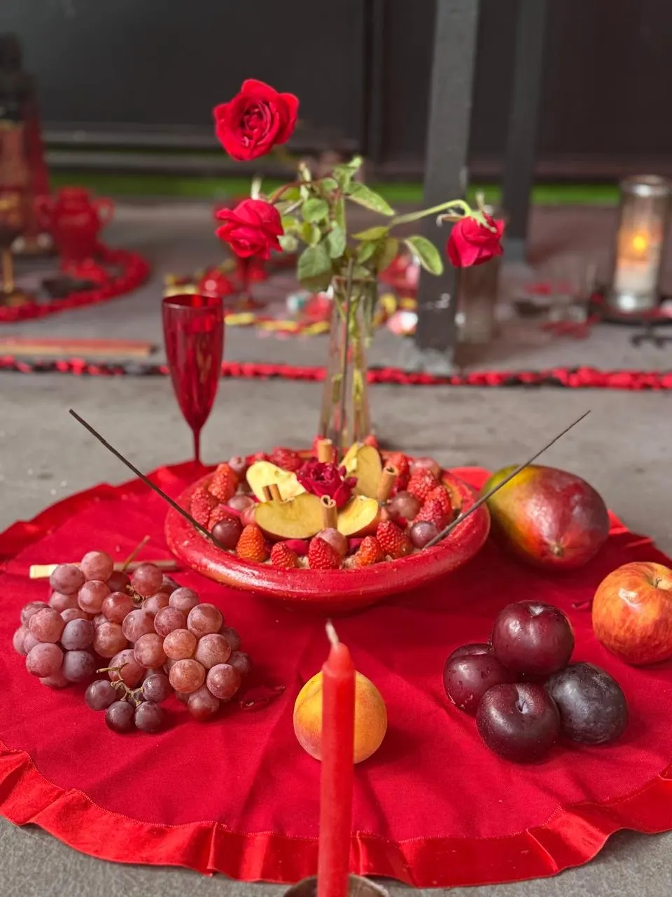 Oferenda sobre toalha vermelha com rosas, uvas, ameixas, maçãs, manga, taça vermelha e um prato de morangos e frutas picadas