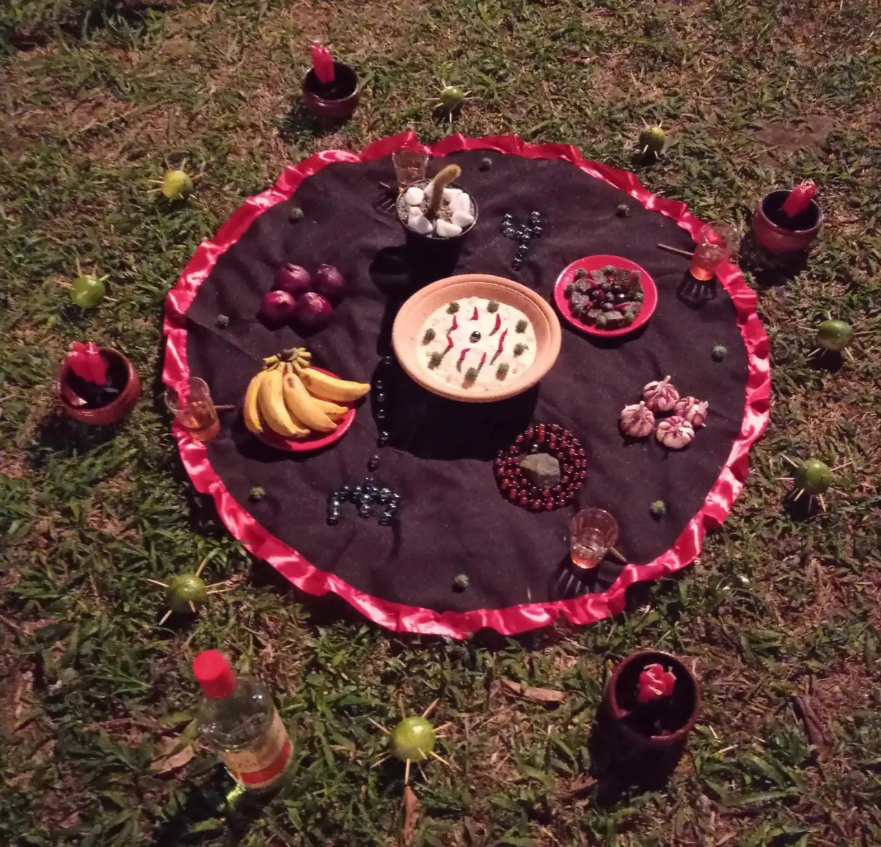 Oferenda a Exu Mirim sobre toalha preta com babado vermelho, no gramado, com bananas, cebolas, frutas e velas em volta