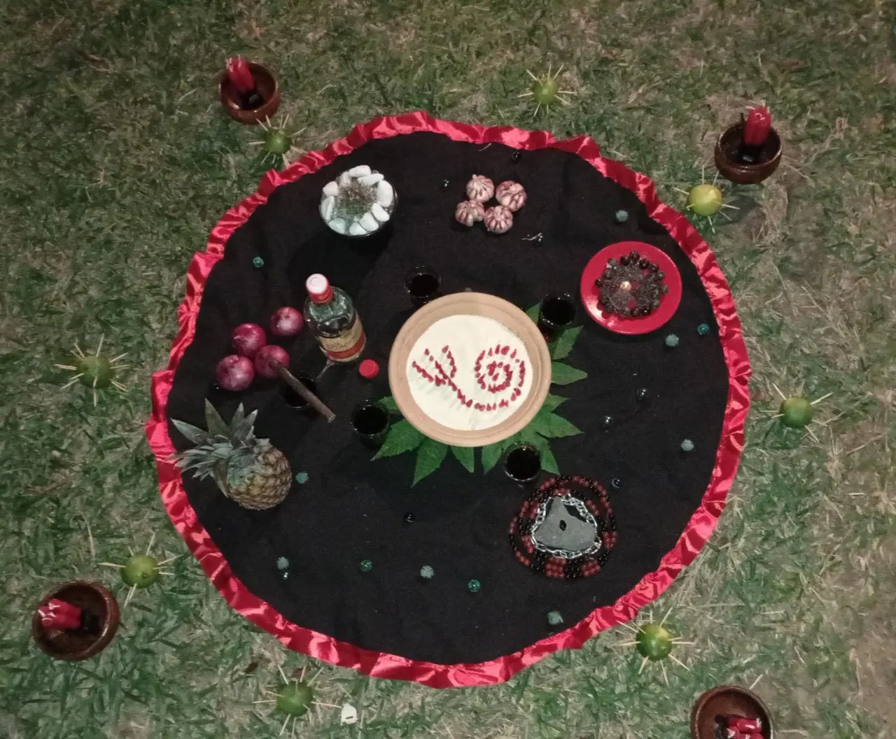 Oferenda a Exu Mirim no gramado, com abacaxi, prato com pimentas em espiral e velas em alguidares