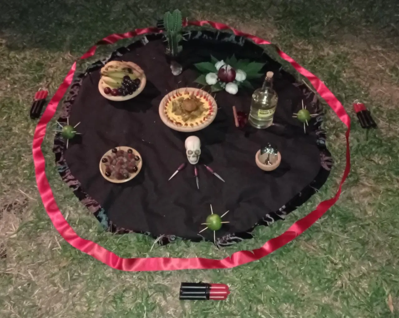 Oferenda sobre toalha preta com fita vermelha em volta, com pratos de frutas, velas, tridentes e uma pequena caveira