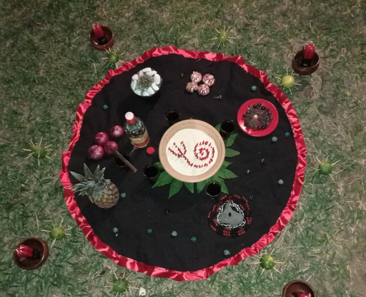 Oferenda vista de cima sobre toalha preta com babado vermelho, no gramado, com prato de farofa ao centro, abacaxi, cebolas roxas, guia de contas e velas em alguidares