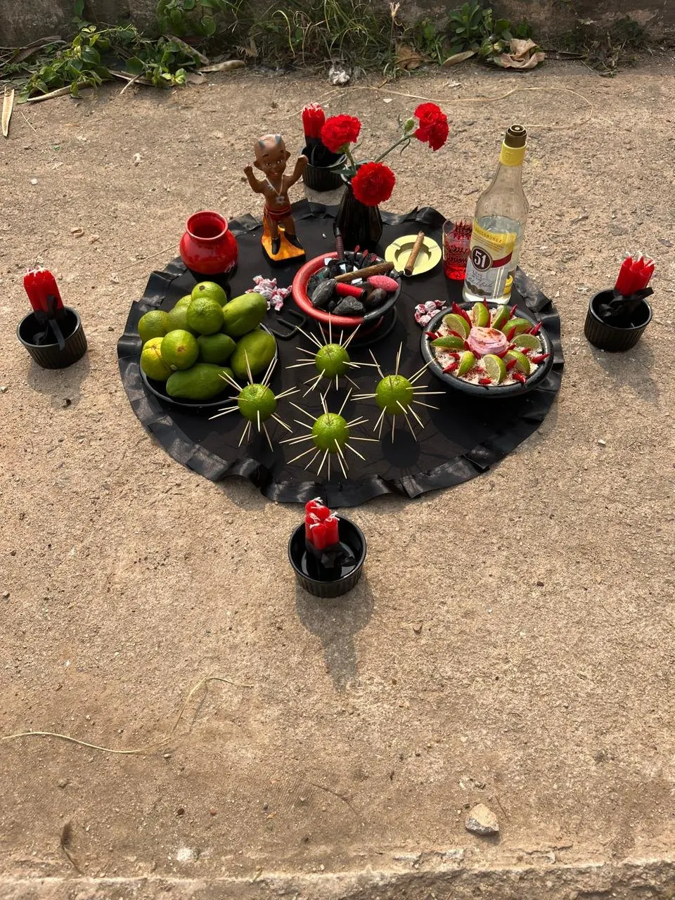 Oferenda sobre toalha preta com limões espetados com palitos, uma pequena imagem, cravos vermelhos, cachaça e velas em alguidares