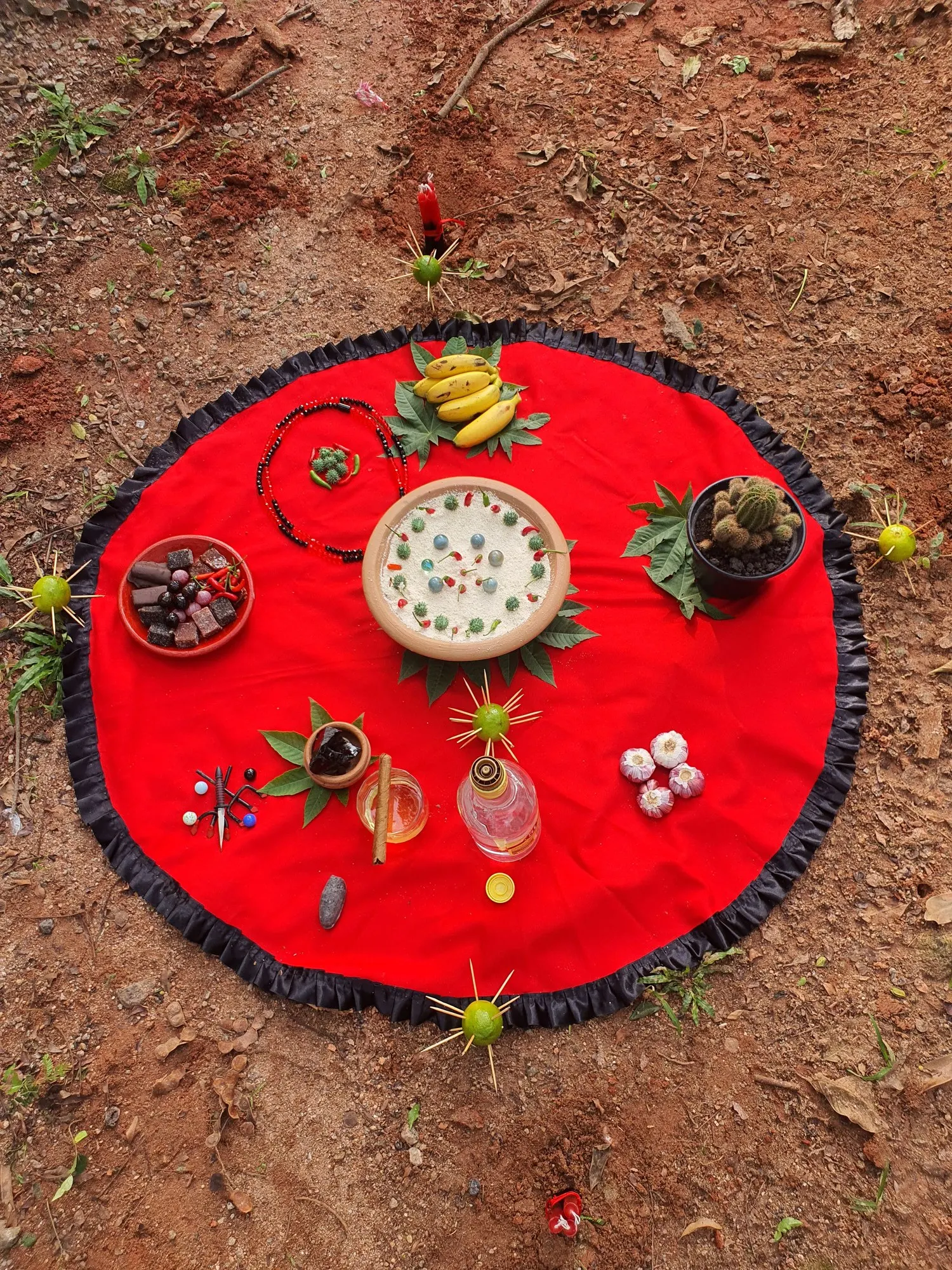 Oferenda sobre toalha vermelha com babado preto, na terra, com frutas e velas