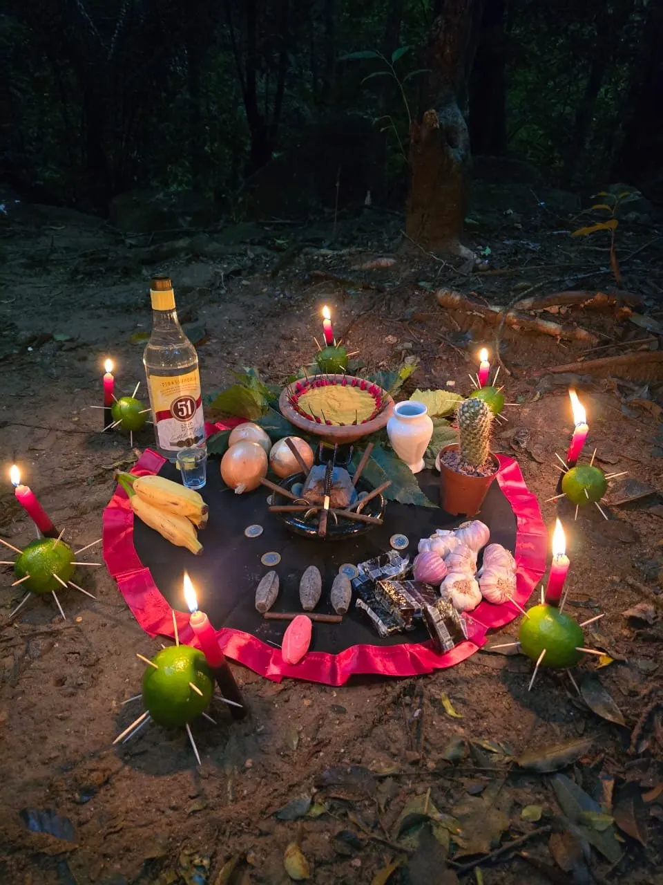 Oferenda na mata, à noite, sobre toalha preta com babado vermelho, com velas acesas, limões espetados com palitos, cachaça, frutas e moedas