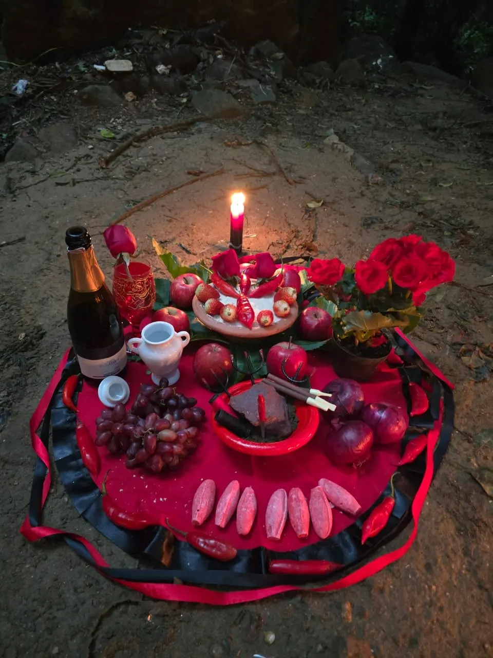 Oferenda à noite sobre toalhas vermelha e preta, com vela acesa, espumante, rosas vermelhas, maçãs, uvas e frutas cortadas