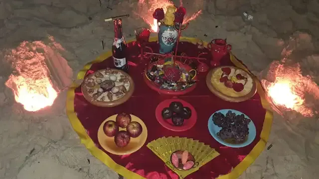 Oferenda na areia, à noite, sobre toalha vermelha e dourada, com frutas, doces e velas
