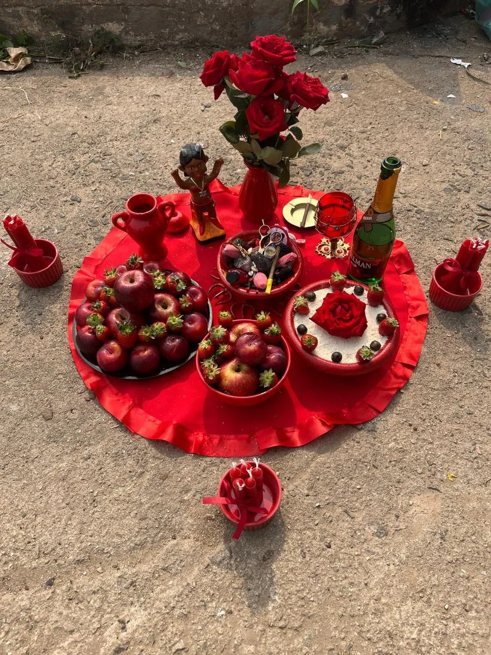 Oferenda sobre toalha vermelha, no chão de cimento, com uma pequena imagem, rosas vermelhas, maçãs, morangos, espumante e velas vermelhas em alguidares