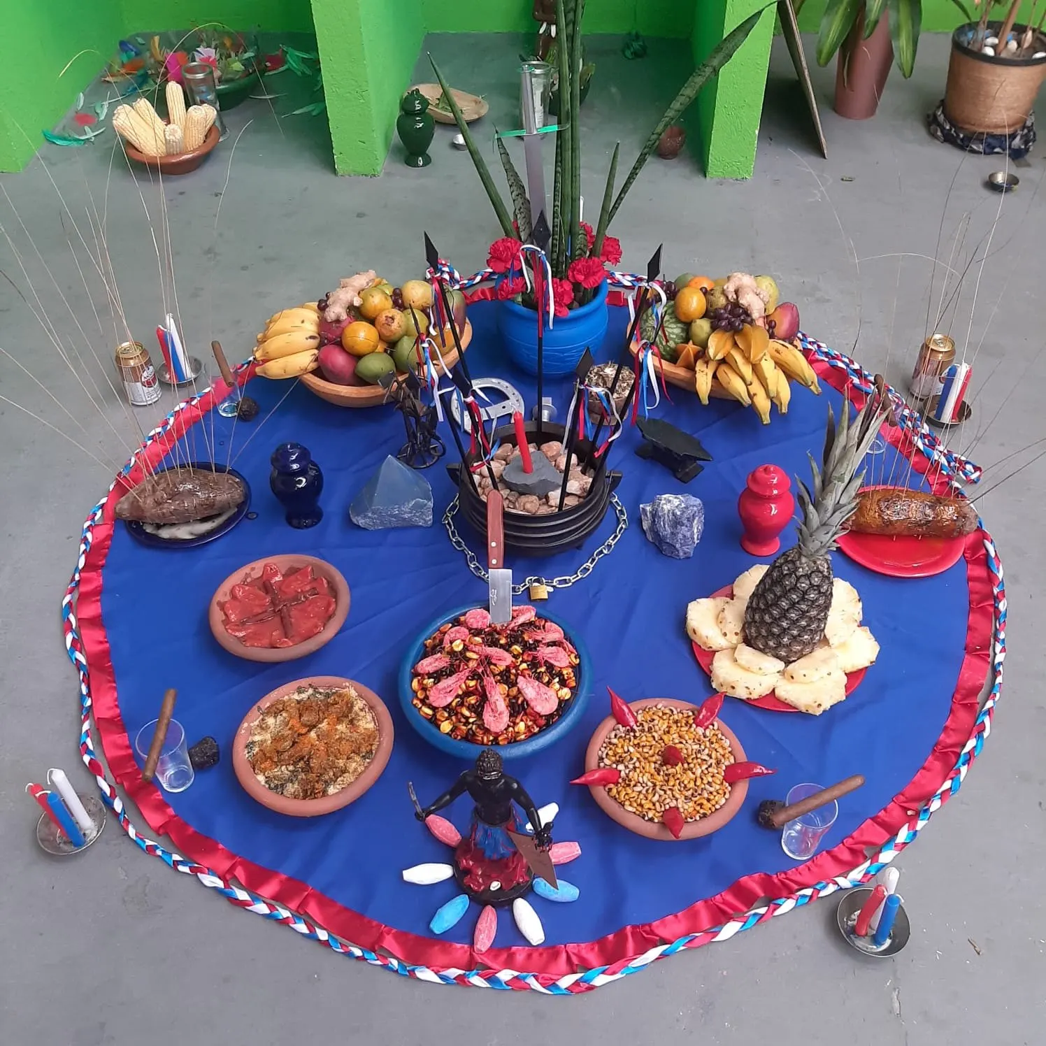 Oferenda no templo sobre toalha azul com fita vermelha e branca, com espadas, corrente, pratos de comida, frutas, abacaxi e flores vermelhas