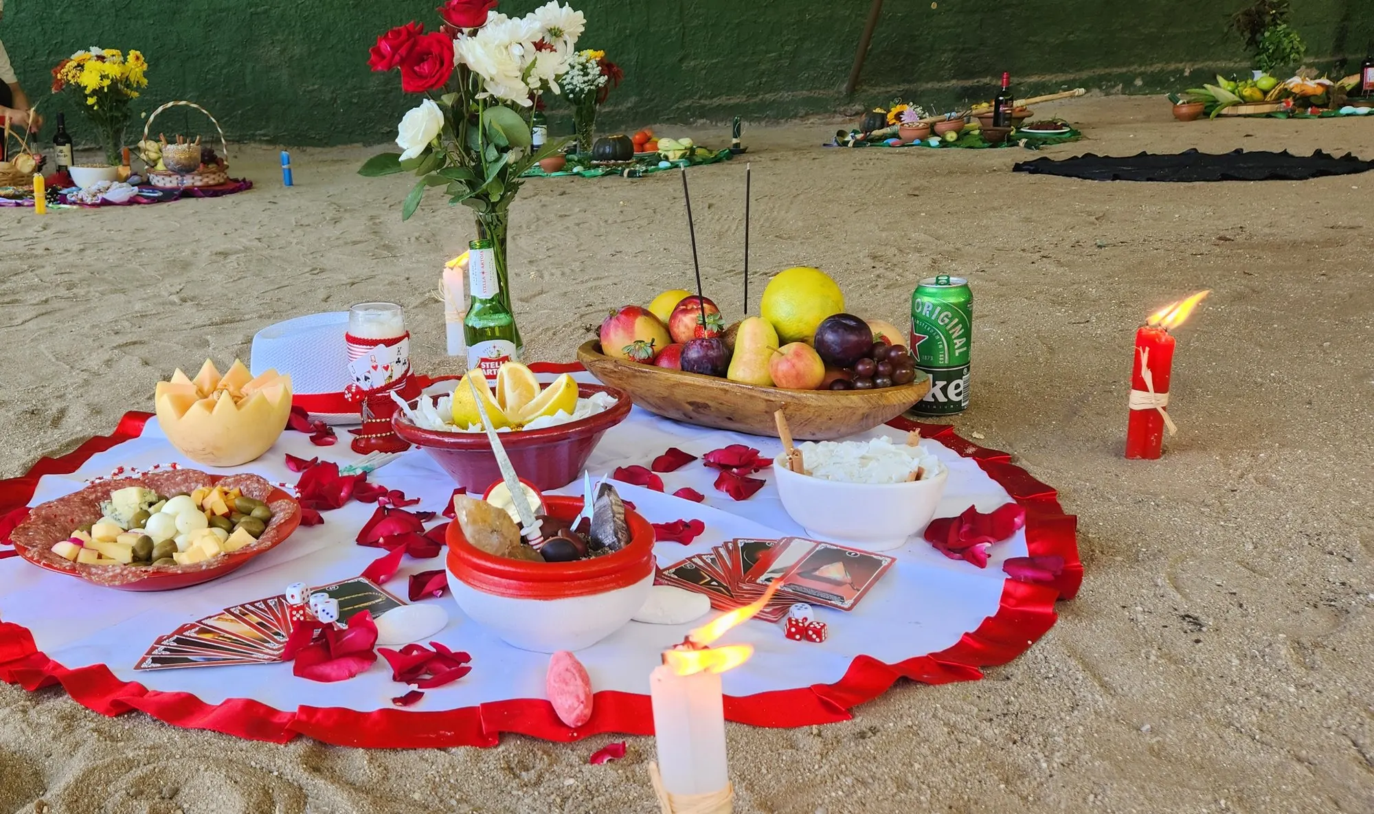 Oferenda na areia sobre toalha branca com babado vermelho, com flores, frutas, pétalas de rosa e velas vermelhas e brancas acesas