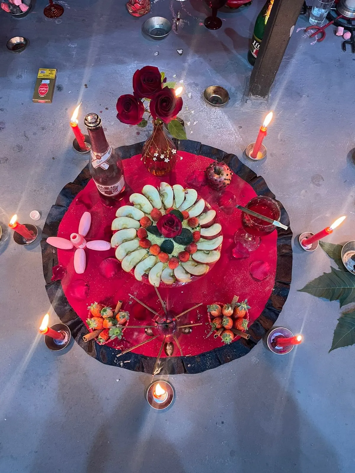 Oferenda vista de cima sobre toalha vermelha com babado preto, com fatias verdes em espiral, morangos, rosas, espumante e velas vermelhas acesas em volta