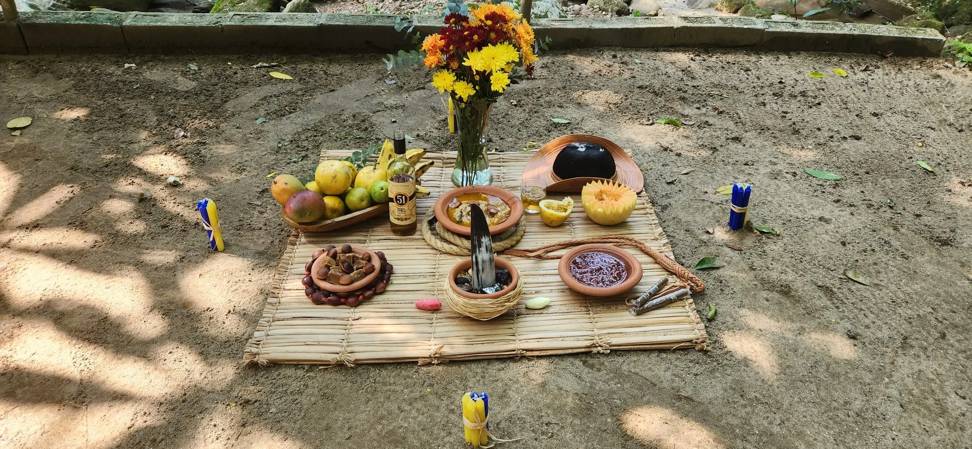 Oferenda sobre uma esteira na terra, à sombra, com flores amarelas e vermelhas, frutas, pratos de barro e velas coloridas