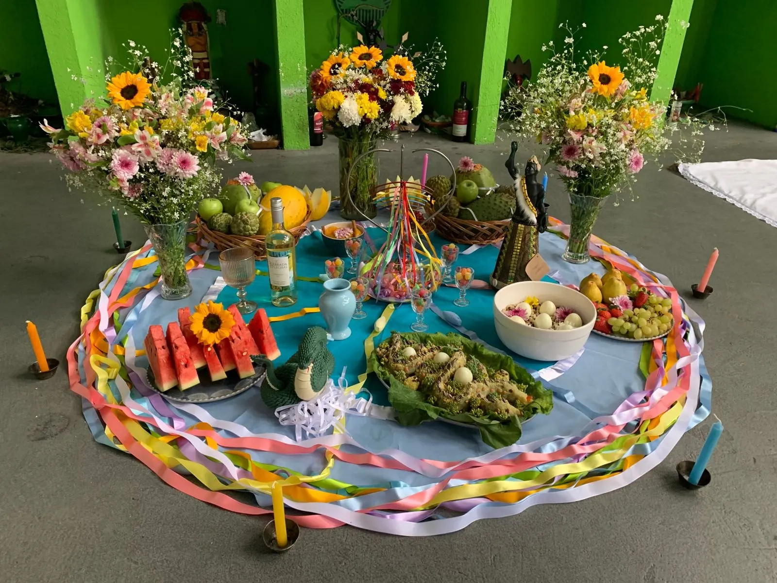 Oferenda no templo sobre toalha azul cercada de fitas coloridas, com girassóis e flores do campo, melancia, frutas, velas coloridas e uma imagem