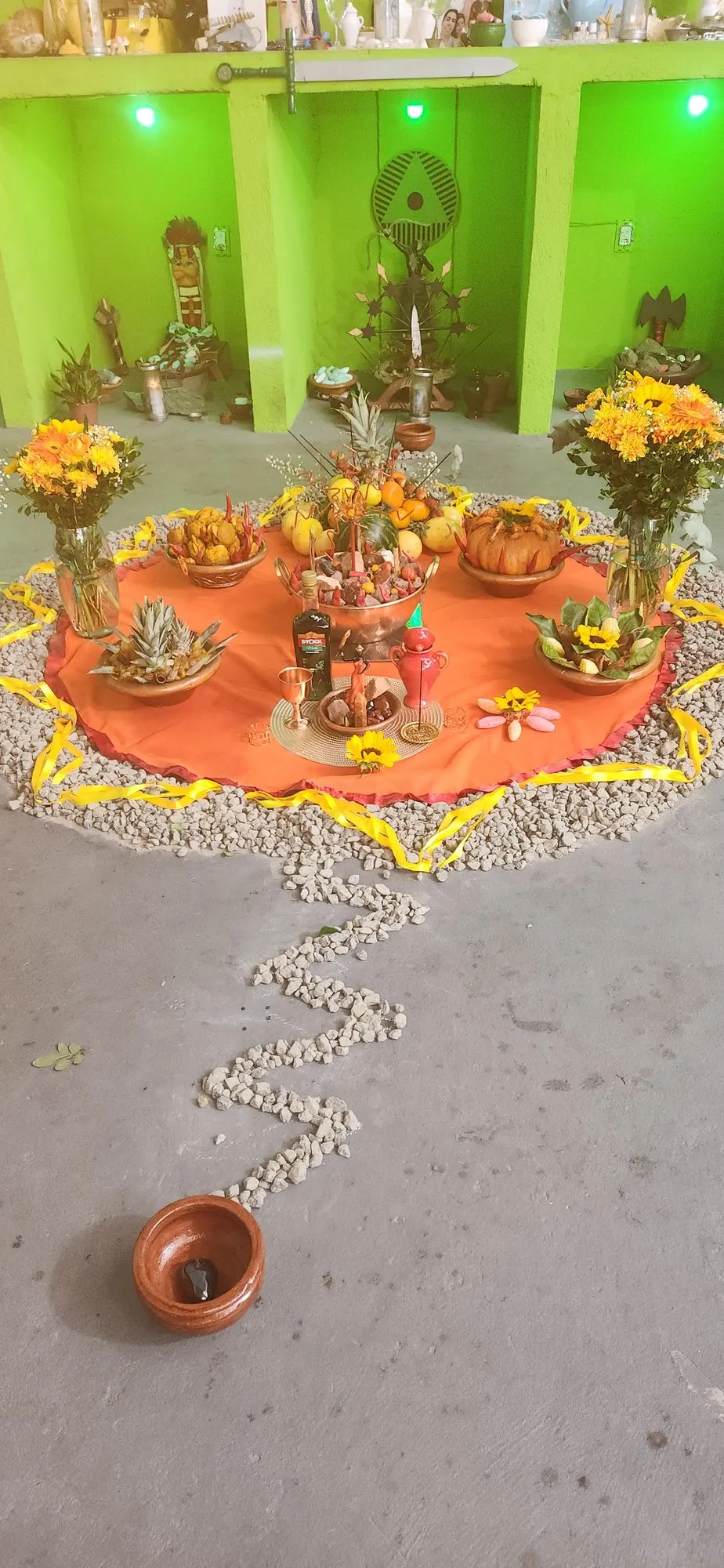 Oferenda no templo sobre toalha laranja, com flores amarelas e laranjas em vasos, frutas e um caminho de pedrinhas brancas até um alguidar