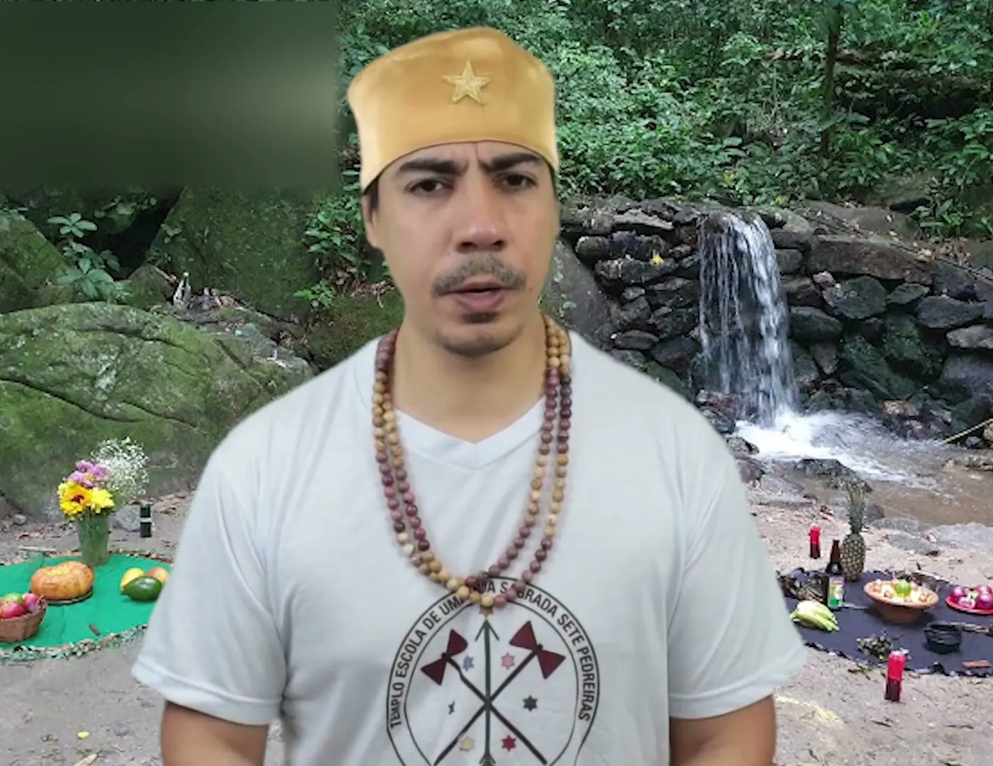 Bruno Amorim, de camiseta branca, diante de uma cachoeira com oferendas no chão