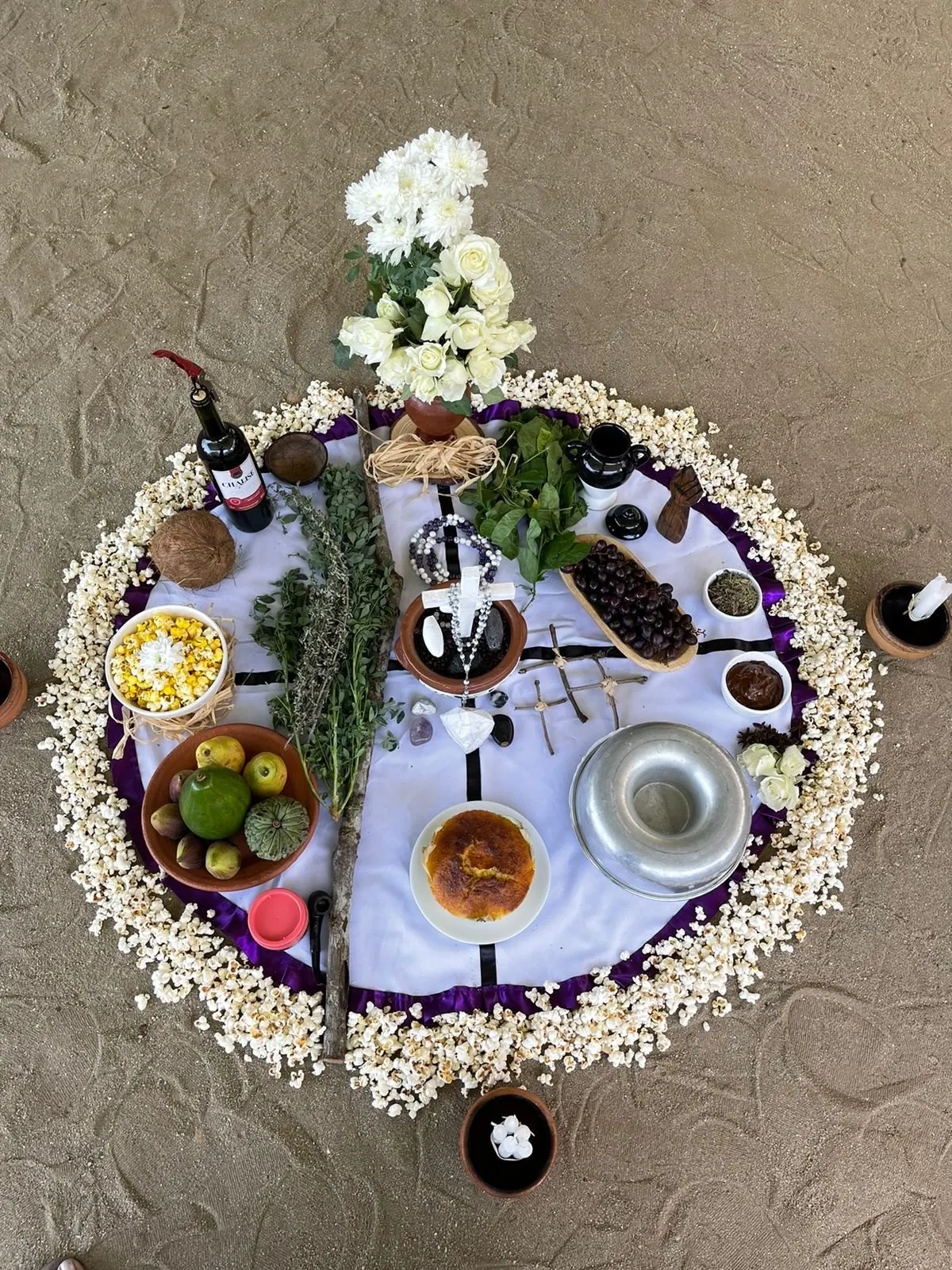 Oferenda vista de cima sobre toalha branca com borda roxa, cercada de pipoca, com flores brancas, ervas, frutas, milho, vinho e velas