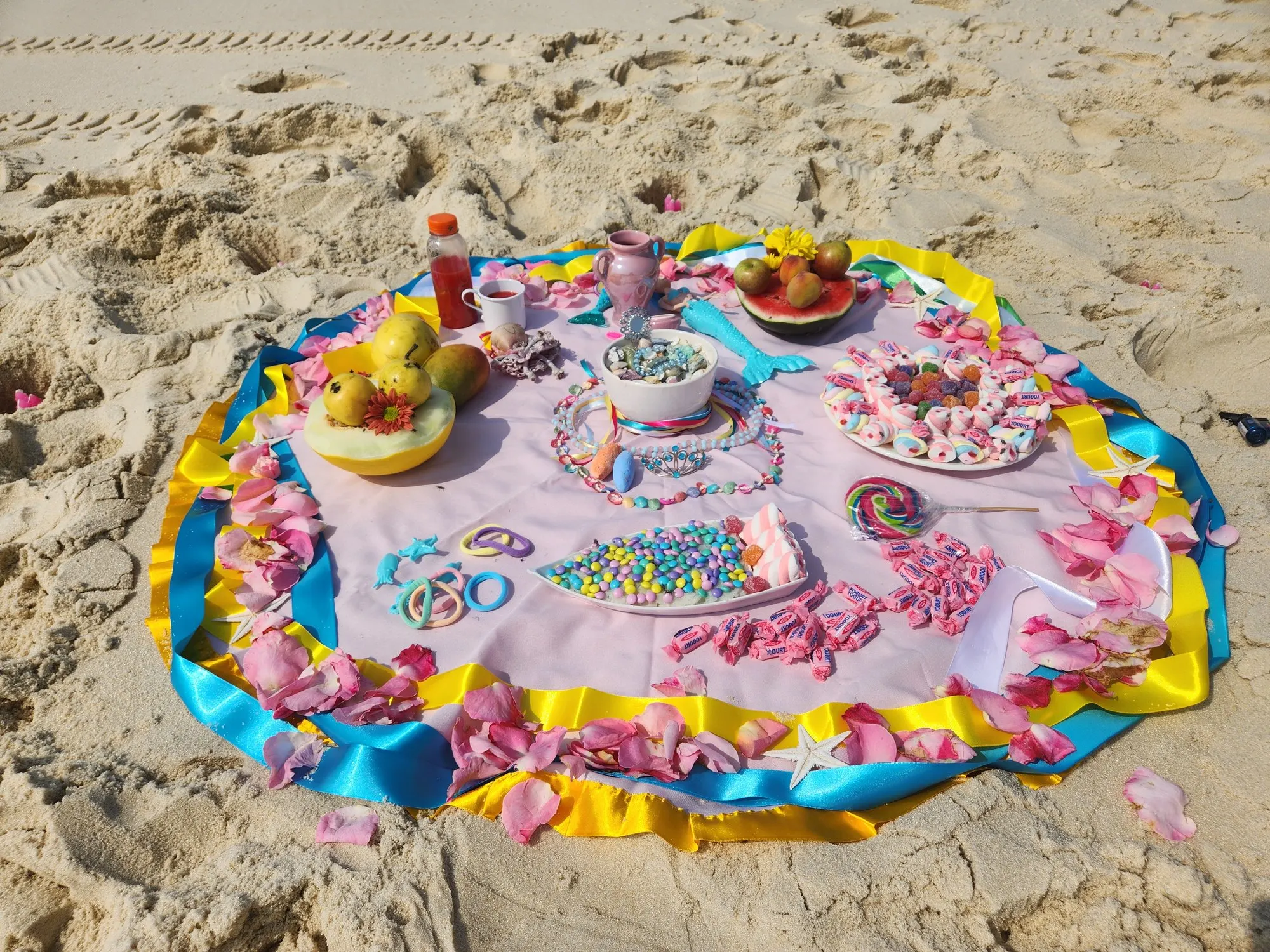 Oferenda na areia da praia sobre toalha clara com fitas azuis e amarelas, com doces, balas, frutas e pétalas cor-de-rosa
