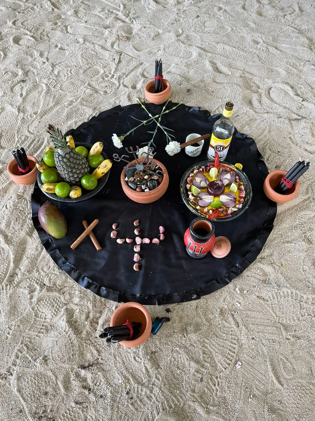 Oferenda na areia sobre toalha preta, com abacaxi, limões, pratos de frutas e pimentas, velas em alguidares e cruzes montadas sobre o pano