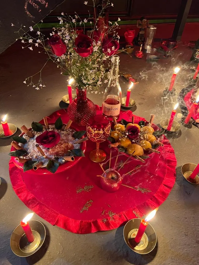 Oferenda sobre toalha vermelha com rosas, flores miúdas, frutas, taça e velas vermelhas acesas em pratos dourados