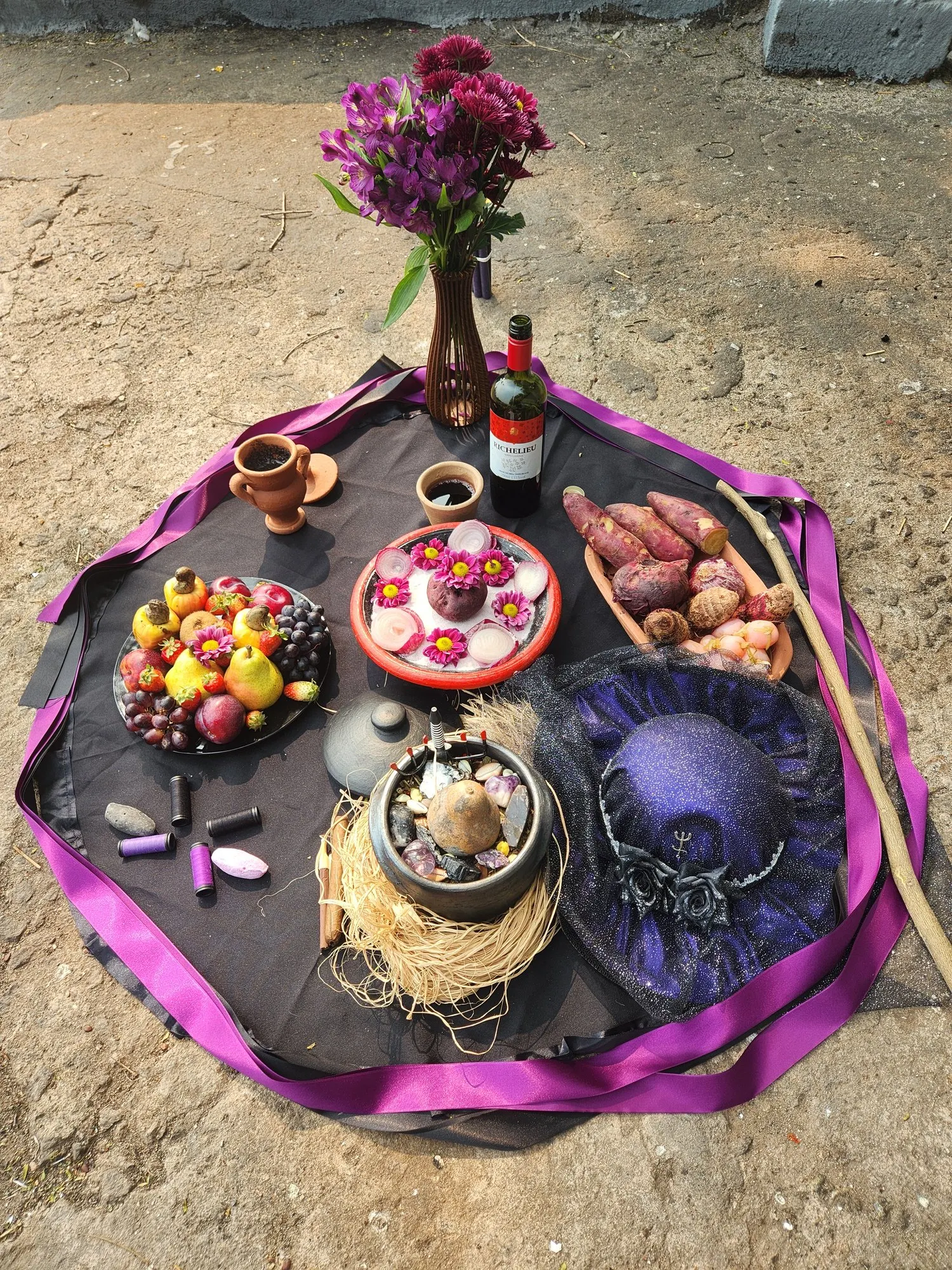 Oferenda sobre toalha preta com fita roxa, com flores roxas, chapéu roxo, frutas, batata-doce, vinho, palha e um cajado