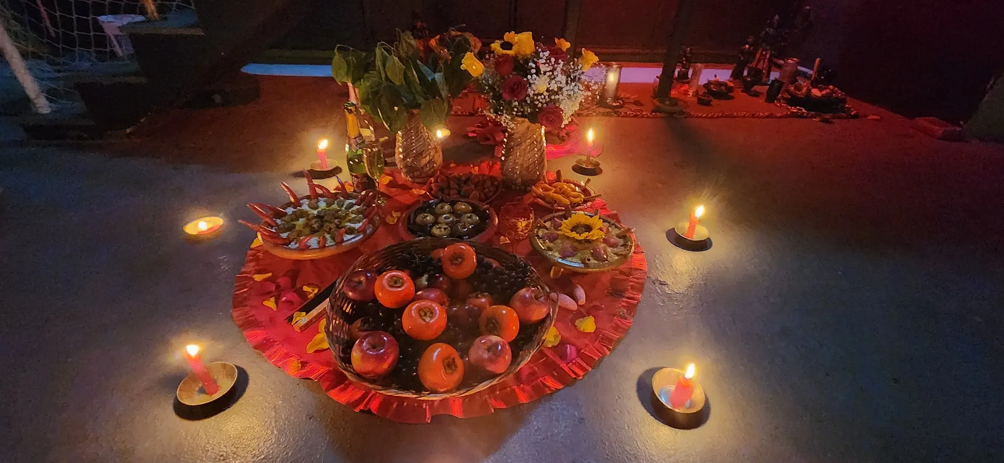 Oferenda à noite sobre toalha vermelha com babado, cheia de frutas e flores, com velas acesas em volta
