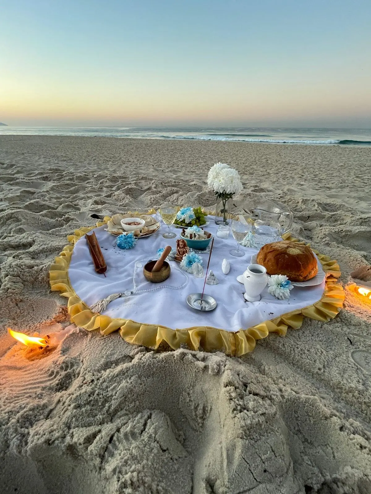 Oferenda na areia da praia sobre toalha branca com babado dourado, com flores brancas, pão, louça branca e incensos, e uma lamparina acesa ao lado
