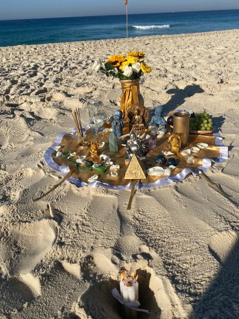 Oferenda na areia, perto do mar, sobre toalha dourada com imagens, uma pirâmide dourada, girassóis, incensos e taças