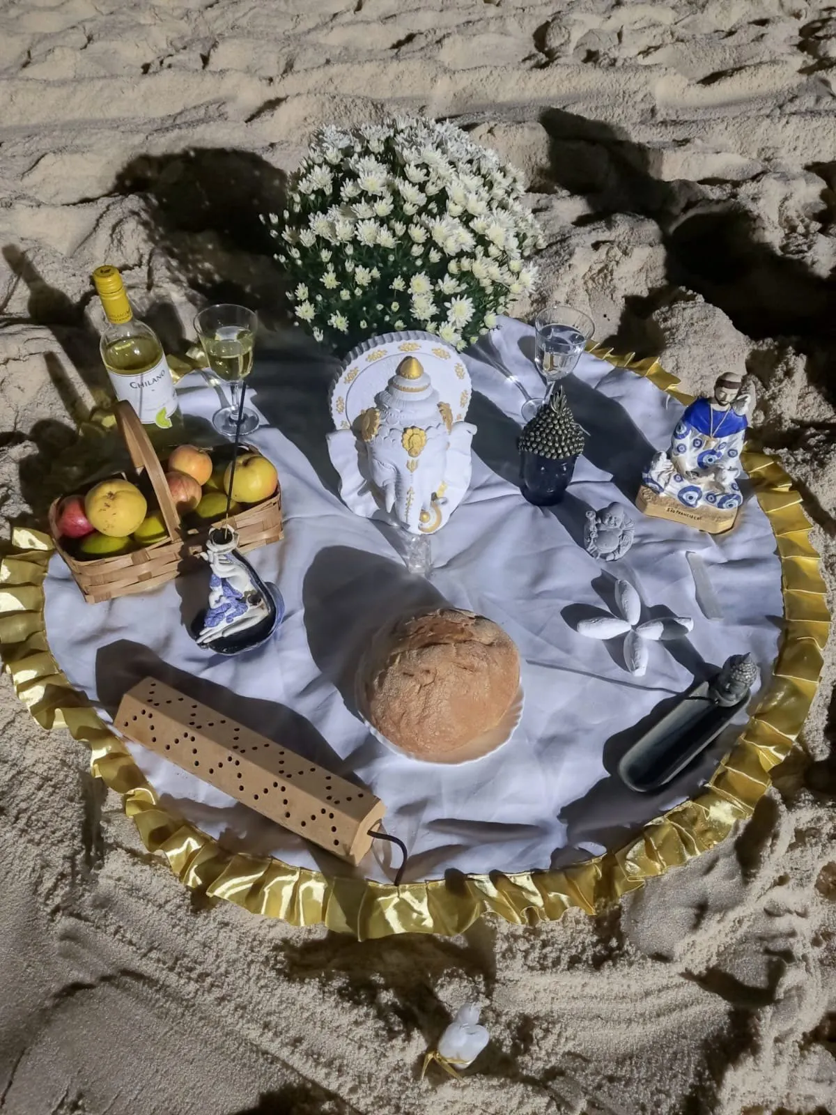 Oferenda na areia, à noite, sobre toalha branca com babado dourado, com crisântemos brancos, pão, cesta de frutas, vinho branco e louças azuis e brancas